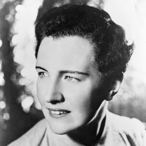Black-and-white close-up photo of Mary Renault, 1946. Her hair is very short and she is wearing a collared shirt. The background is dappled
            with blurred light.
