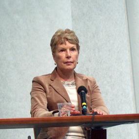 Colour photograph of Ruth Rendell, seated at a table with a microphone and a glass of water. She is wearing a fawn jacket, white shirt, and brown trousers. Her is short and blonde.