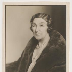 Black and white photograph of Henry Handel Richardson. She is seated, wearing a large fur coat over a white blouse, and her hair is jaw-length, dark, and wavy.