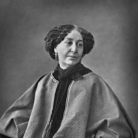 Black and white photograph of George Sand by Nadar, 1864. She is seated, wearing a broad skirt and matching poncho top in thin stripes dress with a dark blouse underneath, a tassel on a long string around her neck, and dangling earrings. Her dark hair, greying a little along the centre parting, is jaw-length and curly.
