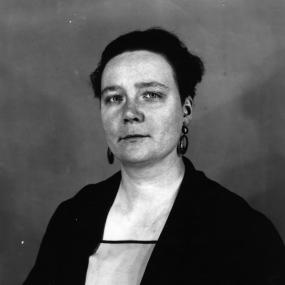 Black-and-white photo of Dorothy L. Sayers. Seated and looking off-camera with a slight smile, she wears large circular earrings and a dark
            blazer. 