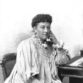 Black and white photograph of Olive Schreiner, seated, with one elbow on a desk and her head resting on that hand. She is wearing a white dress that falls loosely from her shoulders, gathered at the wrists and with ruffles at the throat. Her hair is pinned back with short curls in front.