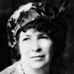 Black and white photograph of Catharine Amy Dawson Scott, shown from the shoulders up. She is wearing a cardigan over a black and white dress, a black beaded necklace, and a puffy hat made of a shiny material, over top of dark curly hair.