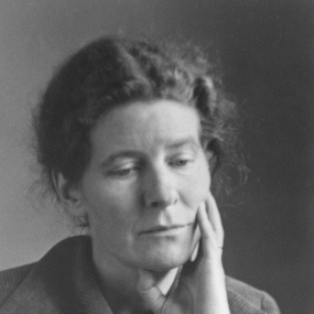 Black and white photograph of E. J. Scovell, shown from the shoulders up. She is wearing a collared corduroy jacket with buttons up the front, and her curly hair is brushed back from a centre parting. Her head is resting on her hand. She looks thoughtful.
