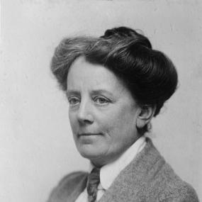 Black and white photograph of Ethel Smyth, shown from the shoulders up. She is wearing a tweed suit jacket with a white shirt underneath and a tie. Her dark is pulled into a bun on the top of her head.