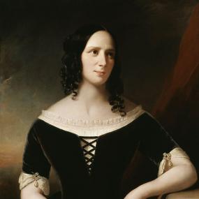 Photograph of a painting of Agnes Strickland by John Hayes, 1846. She is seated with her elbow resting on a wooden table. She is wearing a black velvet dress that laces at the front, over a cream coloured shirt that is trimmed with ruffles. Her sleeves are trimmed with cream coloured lace and pearls, and she is wearing a matching pearl bracelet. Her dark hair is curly, and partly pulled back. She holds a small paper scroll with early-period writing on it, and perhaps a seal. National Portrait Gallery.