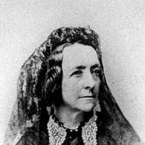 Black and white, head-and-shoulders photograph of Jemima Tautphoeus, wearing a dark dress buttonrf in front and a light lace collar. She has dark, chin-length, wavy hair, and a shawl over her head and shoulders.