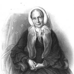 Black and white photograph of a drawing of Ann Martin Taylor, seated in a chair that is upholstered with flowers, wearing a simple, long, dark dress with a high collar and wide sleeves. She is wearing a white bonnet that is tied in a bow at her chin and a pair of small wire spectacles. Signed underneath the drawing are the words: affectionately yours, Ann Gilbert.