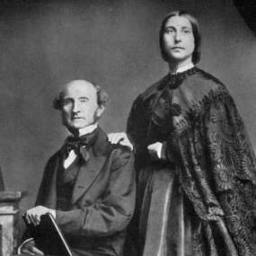 Black and white photograph of Helen Taylor, standing with her hand on the shoulder of her seated stepfather, John Stuart Mill.  She is wearing a long, simple dark dress with a wide skirt, and a dark embroidered shawl. Her dark hair is pulled back and tied at the nape of her neck.