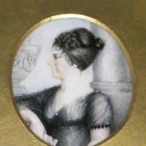 Photograph of a drawing of Anna Jane Vardill, shown from the waist up, wearing a black dress that crosses at the bust, and has short puffed sleeves. Her dark hair is pulled back, with braids wrapped around the front and curls at the top. Behind her is a large column and a vase carved with people's faces displayed on a pedestal. The circular photograph is contained within a gold frame, and the name "Mrs. Niven" is written underneath it.