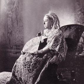 Black and white photograph of Queen Victoria, done for her Diamond Jubilee, 1897, by W. and D. Downey. She is seated, face in profile, holding a fan and wearing an elaborately embroidered floor-length dress, a small crown, a veil on the back of her head whose lace matches that of her skirt, a jewelled bracelet and drop earrings. National Archives of Canada.