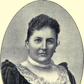 Black and white oval photograph of Lucy Walford, set against a light yellow background. She is wearing a dark, long-sleeved dress with a touch of lace at the neckline. Her dark curly hair is pulled back.