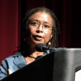 Photograph of Alice Walker, standing behind a podium and speaking into a microphone. She is wearing a blue jacket, dangling leaf-shaped earrings, and wire-rimmed glasses.