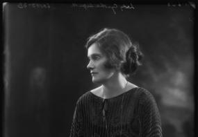 Black and white photograph of Lady Cynthia Asquith, in profile, sitting on a carved bench. She is wearing a loose-fitting dress with thin stripes and a pattern superimposed, a wrist-watch, and a long, simple pendant. Her hair is loosey pinned back in a bun.