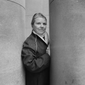 Black-and-white photograph of Joan Aiken, who is standing between two large columns, 19 October 1984. 