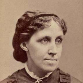 Sepia head-and-shoulders photograph of Louisa May Alcott by Warren's Portraits, c. 1870.  She looks pensively away from the camera, wearing a dark dress with two frills up the front, her wavy hair tied back in a bun at the nape of her neck.