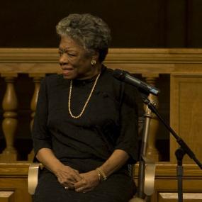 Colour photo of Maya Angelou speaking at Wake Forest University in Winston-Salem, North Carolina. She is sitting, talking away from the microphone on her right, wearing a black dress with gold chain and disc earrings, hands clasped in her lap.