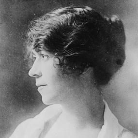 Black and white, head and shoulders photograph of the adult Daisy Ashford in 1919. She is seen in profile, with her dark hair in a bun, wearing a light gauzy dress with a brooch at the centre of the neckline.  She has signed the photo with her name and "24.11.19."
