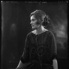 Black and white photograph of Lady Cynthia Asquith, in profile, sitting on a carved bench. She is wearing a loose-fitting dress with thin stripes and a pattern superimposed, a wrist-watch, and a long, simple pendant. Her hair is loosey pinned back in a bun.