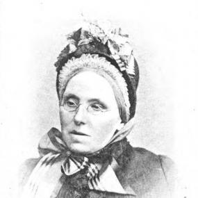 Black and white, head-and-shoulders photograph of Isabella Banks by Done and Ball of London. She has her head slightly tilted under a hat with frill edges and bows for ornament, with a large one under her chin holding it on. She has grey hair and thin wire-framed glasses.