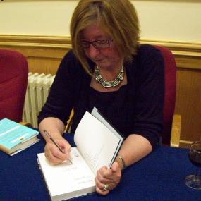 Colour photo of Pat Barker signing a copy of her book "Toby's Room". She sits behind a table with a glass of red wine, wearing a black top and silver necklace.