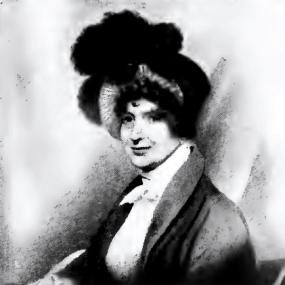Black and white photo of a miniature of Lady Anne Barnard. She sits writing with a quill on a sheet of paper, but pauses to look up and to her right with a half-smile. She wears a loose open jacket over a white high-necked blouse, and a large, soft hat in several dark and white layers, with dark feathers on top. Her hair, too, is dark.