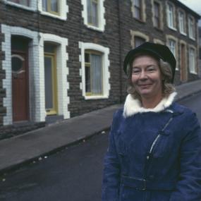 Photo of Nina Bawden in front of a row of small houses with stone facades, wearing a blue coat and a black hat, 1974. 