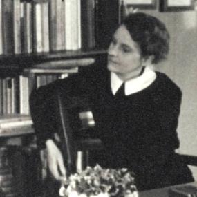 Black and white photograph of Sylvia Beach, seated at a desk, with a bookshelf behind her. She is turned, looking off to the side, and she is wearing a plain black dress with a white collar.
