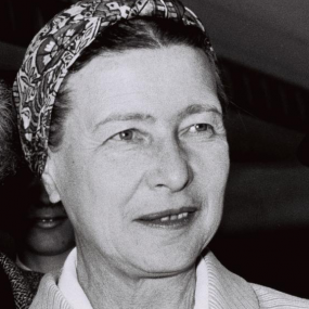 Black and white, head-and shoulders photo of Simone de Beauvoir (with someone else just visible behind her). She is wearing a heavy top with stripes over a white shirt. Her hair is mostly hidden by a patterned scarf worn turban-style.