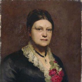 Painting of Lydia Becker by Elizabeth Sarah Guinness. Becker is wearing a black dress with white lace collar and a bunch of red flowers. She has a pendant hanging on a black strip, and wire-framed glasses. Her hair is parted in the middle and scraped back in a bun. Girton College, Cambridge.