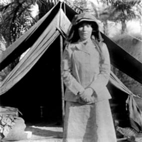 Black and white photo of Gertrude Bell standing in front of a tent in Iraq, 1941, palm trees in the background. She has a covering on her head to protect her from the sun. She is wearing a jacker, a long skirt, and unsuitable-looking shoes.
