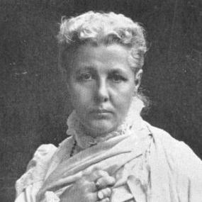 Photograph of Annie Besant looking directly at the camera with her right hand over her chest. She has a serious expression and is wearing a white high frill-collared dress with possibly a shawl over it. She has two rings on her visible right hand. She has blonde hair and it is in an updo.