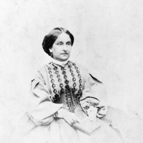 Black and white photograph of Isa Blagden, seated with a book in her lap. She wears a dress with wide skirt, puff sleeves, a high collar, a pattern of stitched swirls going up the torso, and a tightly laced waist covering like a corset. Her dark hair is pinned at the back of her head.