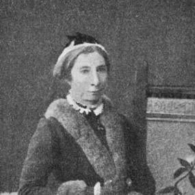 Black and white photograph of Jessie Boucherett, standing with one arm resting on the back of a chair. There is a screen with a pattern of vines behind her, and she is wearing a fur-trimmed coat and a small hat.