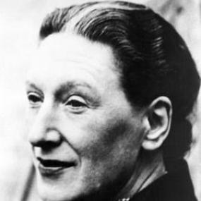 Black and white head-and-shoulders photograph of Elizabeth Bowen seen in profile, with her head slightly turned. She is wearing a dark, high-collared jacket with a polka-dotted scarf around her neck; her hair, dark with a white streak, is pulled back in a bun, and she is wearing dark lipstick.