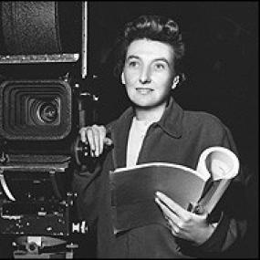 Black and white photograph of Muriel Box as film director, looking alertly forward with one hand on a large camera and the other holding an open script. Her dark, zippered jacket is open over a white shirt.