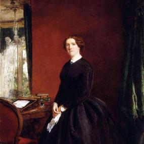 Painting of Mary Elizabeth Braddon by William Powell Frith, exhibited 1865. She stands beside a desk with writing materials and stacks of books, looking at the viewer, in a long dark dress. The wall behind her and the desk chair are red. Her red-brown hair is coiled over her ears, and her clasped hands hold a handkerchief. National Portrait Gallery.
