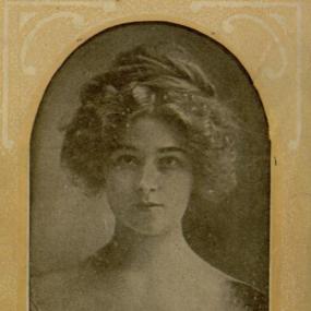 Image of a black and white head-and-shoulders photograph of Charlotte Mary Brame, From a simple brown frame, she stares straight at the viewer, wearing a delicate lacy dress, her hair mostly forming a crown around her head, with a frizz of curls framing her face.