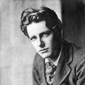 Black-and-white, head-and-shoulders photo of Rupert Brooke taken by Sherril Schell in 1913. Brooke leans a little forward and turns to look into the camera. He has wavy hair, and is wearing a suit and large cravat.