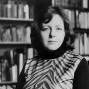Black and white photograph of Brigid Brophy wearing a zebra print sleeveless blouse over a black turtle neck. In the background, there are several bookcases visible. She is facing the camera.