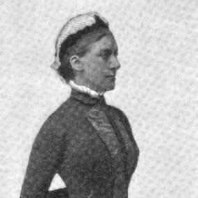 Photo of Rhoda Broughton standing very upright, facing a 3/4 turn away from the camera towards our right. She is wearing a long black, quilted dress and a white cap.