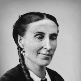 Black and white, head-and-shoulders photograph of Frances Browne, shown at a three quarter turn. She is wearing a dark jacket with a white collar outside it, and large silver hoop earrings. Her straight hair is pulled back and hangs in a long plait down one side.
