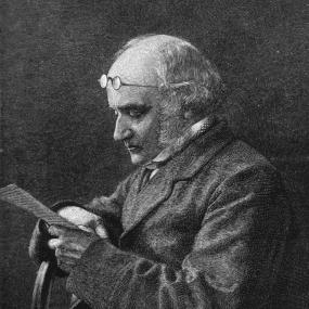 Black-and-white, half-length photo of John Brown, in profile, seated; he reads from a paper, with his glasses pushed up on his head. He is clean-shaven below full sideburns.