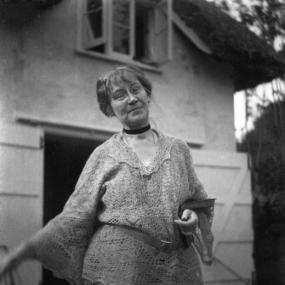 Black and white photo of Dorothy Bussy outside a house. She is wearing loose-fitting trousers and a loose lace-patterned shirt belted at the waist. She has small wire-rimmed spectacles, a simple black ribbon around her neck, and short hair with a fringe. She has her head slightly on one side, an open book tucked under her arm, and a slight, quizzical smile.