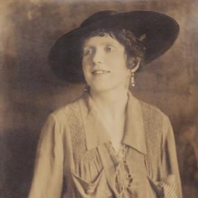 Sepia photograph of Mary Butts, smiling, seated with her head turned to the side. She is wearing a dark, wide-brimmed hat and a loose-fitting shirt with a smocked yoke and pockets on each side, fastened at the neck with lacing. Her dark hair is short and curly, worn with dangling earrings.