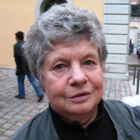 Colour photograph of A.S. Byatt, shown from the shoulders up. She is wearing a black fleece sweater with a high neck, zipped almost to the top, with a grey cardigan over top. Her hair is short and grey and she stands against the background of a street.