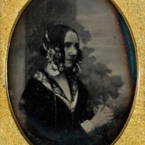 This image is of a daguerreotype of Ada Byron, taken by Antoine Claudet in about 1843. She wears a black dress with lace detail along the neckline and cuffs, and elaborate hair ornament. She sits with clasped hands at a three-quarter angle to the viewer's right. The trees behind are a painted backdrop.