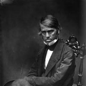 Black-and-white, three-quarter-length photograph of Thomas Carlyle,  taken by Robert Scott Tait on 31 July 1854. Carlyle sits on a carved wooden chair, looking down with a rather grim expression. He has thick straight hair, and has his arms crossed, with a large hat on his knee.