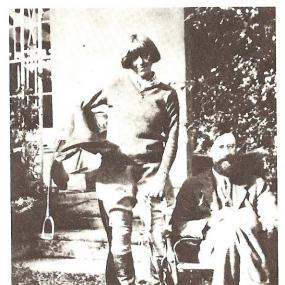 Black and white photo of Dora Carrington standing beside a seated Lytton Strachey in a garden. She is wearing jodhpurs, riding boots, and a sweater over a shirt. Her hair is bobbed and she holds a horse's saddle with the left stirrup hanging down.
