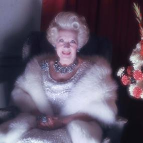 Photograph of Barbra Cartland, seated in a silver upholstered armchair, with her hands clasped in her lap. She is wearing a silver dress with sparkly beading, a white fur shawl, and a large diamond necklace and earrings. Her hair is short, white, and styled in large curls. There is a red curtain behind her and a bouquet of pink and white flowers at the edge of the photograph's frame.